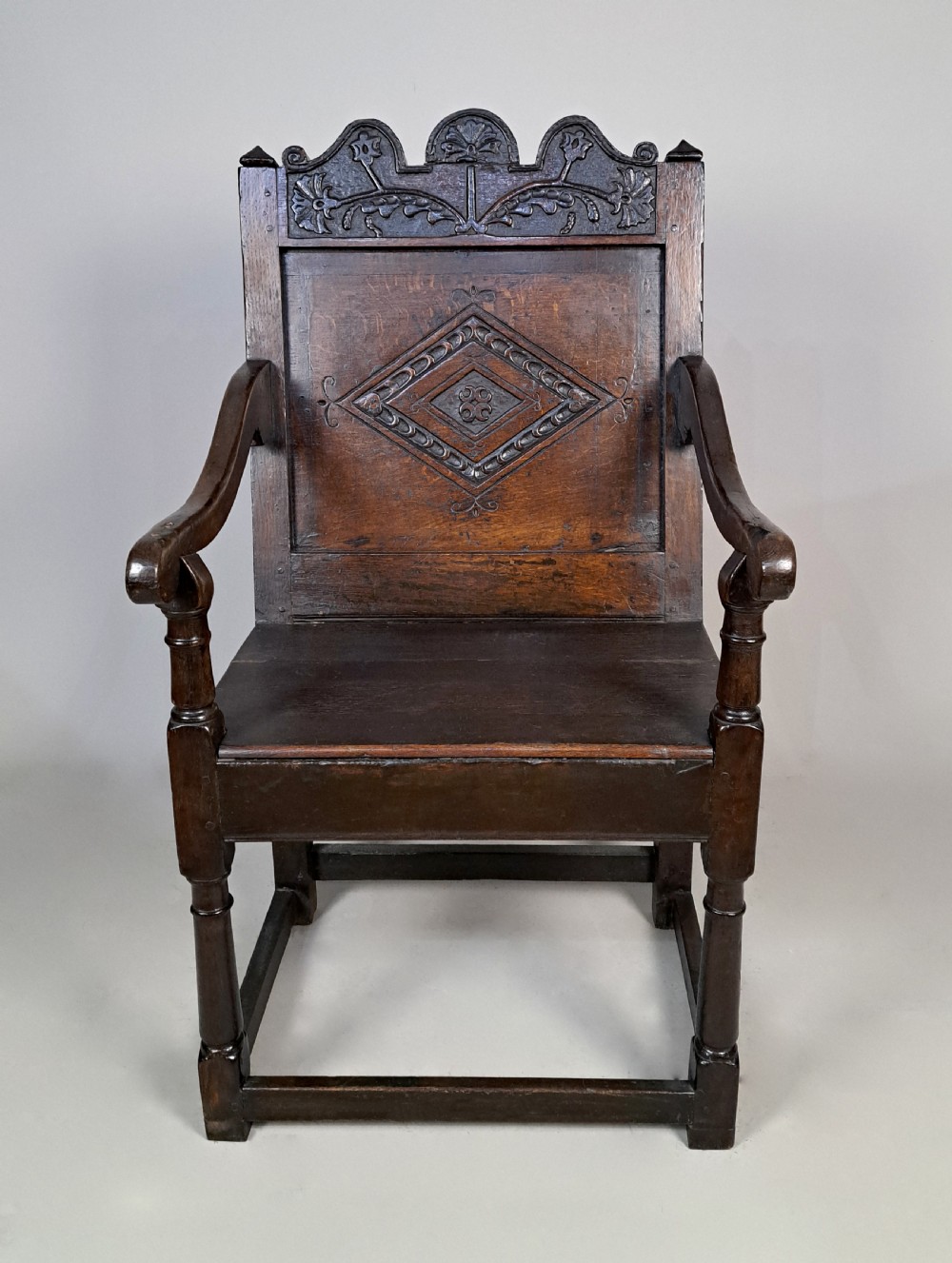 A dark oak wainscot armchair featuring a carved lozenge motif on the back panel, shaped cresting rail, shaped armrests, and turned front legs with lower stretchers.