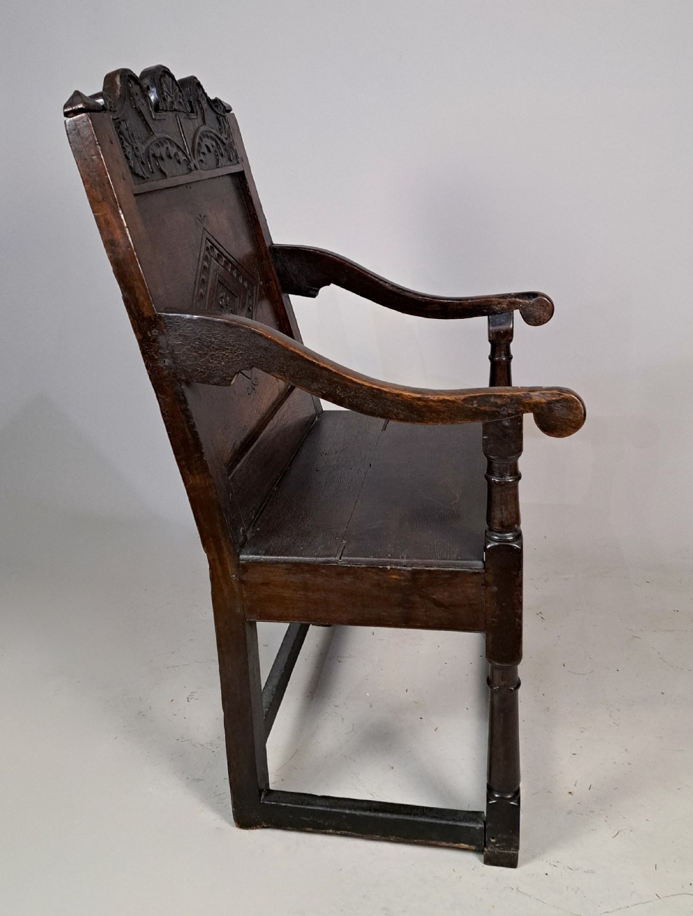 A dark oak wainscot armchair featuring a carved lozenge motif on the back panel, shaped cresting rail, shaped armrests, and turned front legs with lower stretchers. View VI