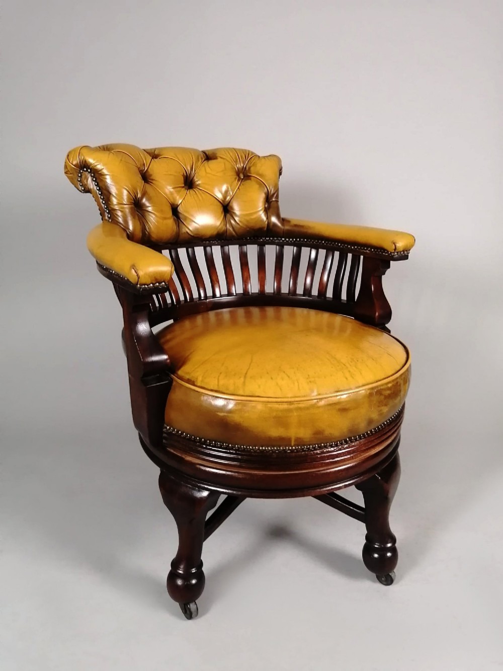A late Victorian revolving desk chair featuring mustard tan buttoned leather upholstery, brass studding, a mahogany spindle-back frame, and turned legs on castors. View IV
