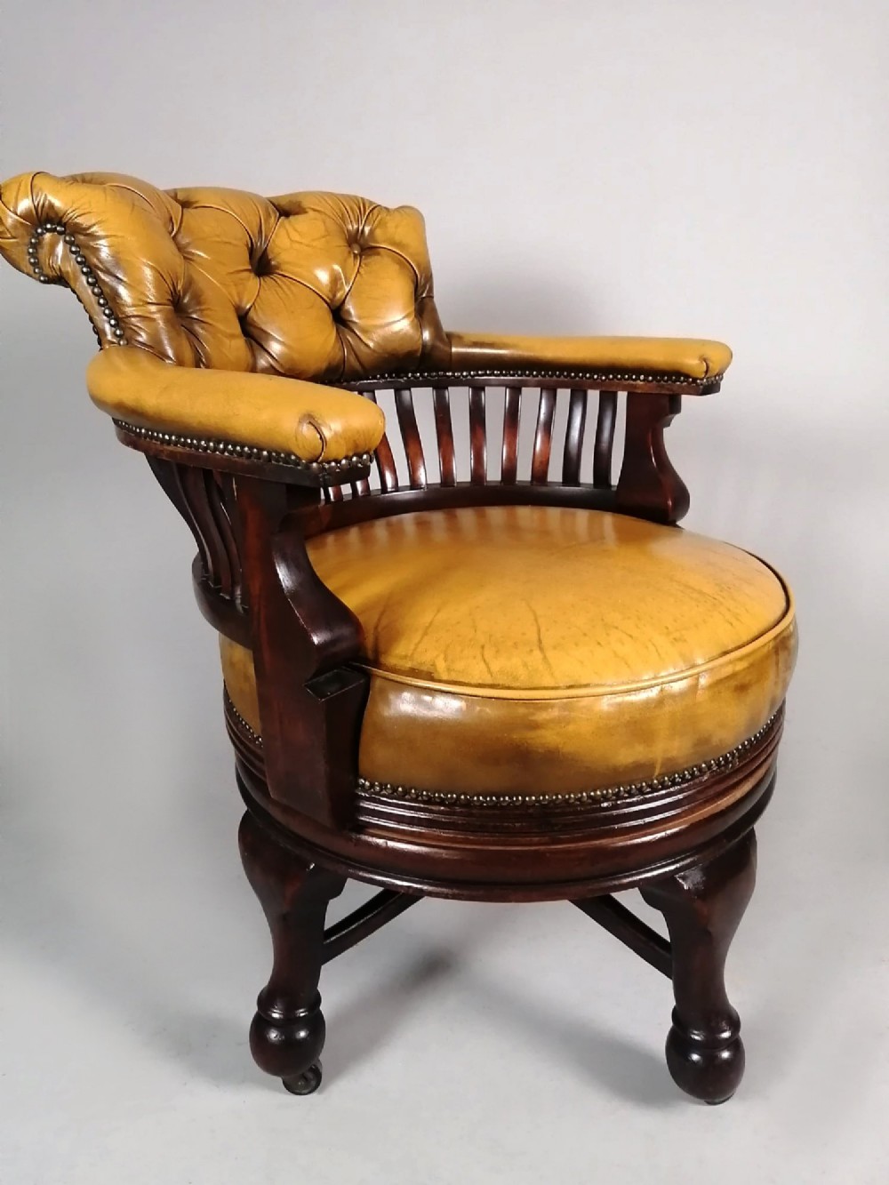 A late Victorian revolving desk chair featuring mustard tan buttoned leather upholstery, brass studding, a mahogany spindle-back frame, and turned legs on castors. View V