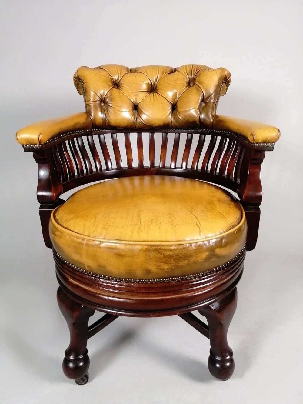 A late Victorian revolving desk chair featuring mustard tan buttoned leather upholstery, brass studding, a mahogany spindle-back frame, and turned legs on castors. View VI