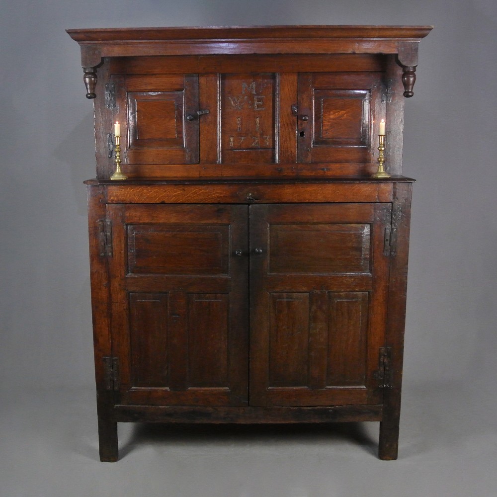 An 18th-century dark oak court cupboard featuring a carved date of 1723, upper recessed cabinet with drop finials, and fielded lower door panels.