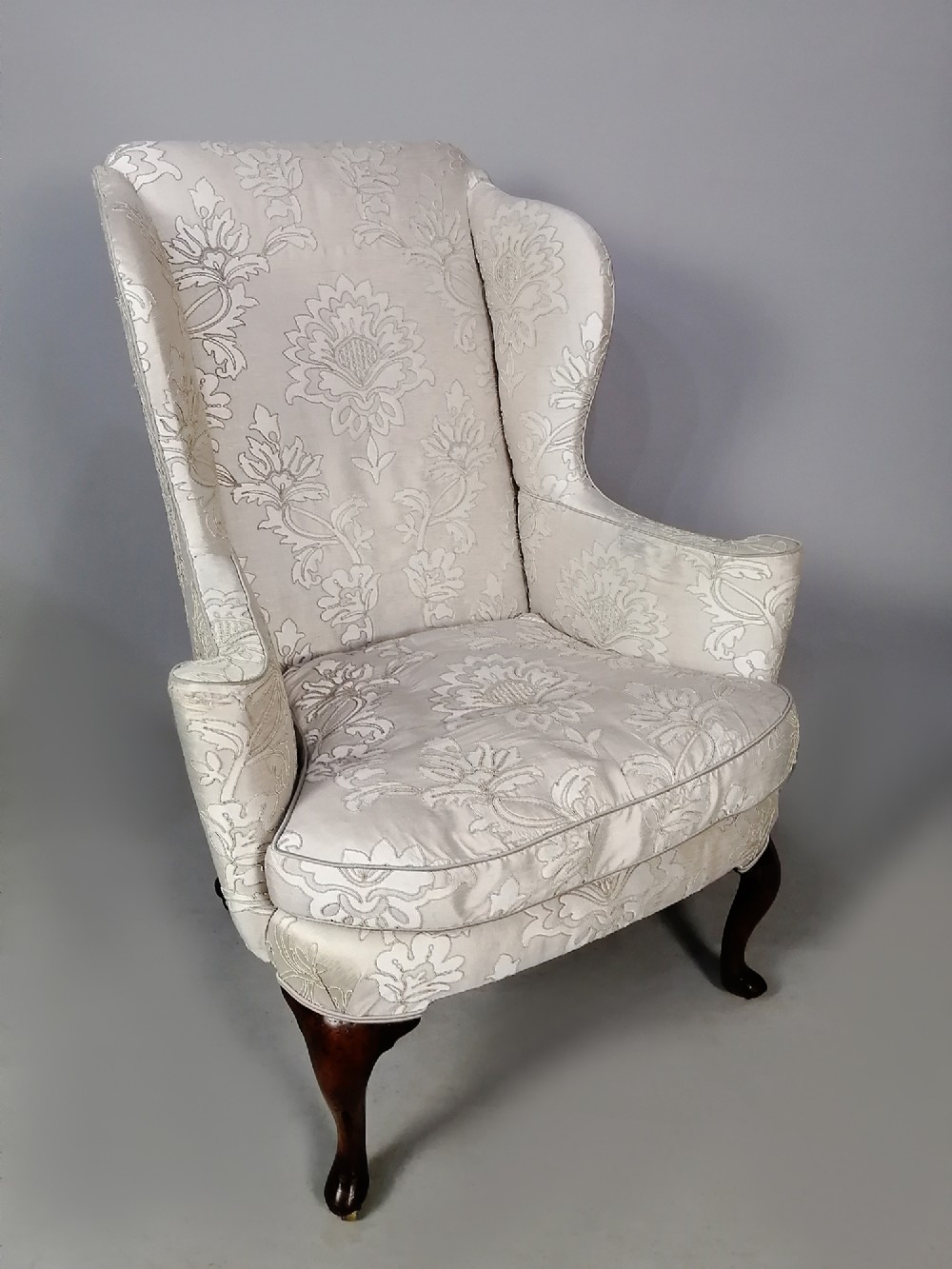 A 19th-century English wingback armchair upholstered in cream damask fabric with dark carved walnut cabriole front legs.