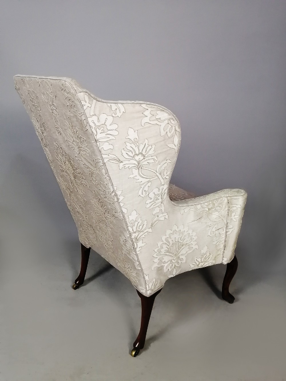A 19th-century English wingback armchair upholstered in cream damask fabric with dark carved walnut cabriole front legs. View V