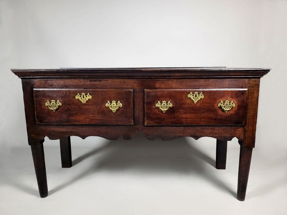 A George III dark oak dresser base featuring two drawers with ornate brass handles and escutcheons, a shaped apron, and tapered legs.