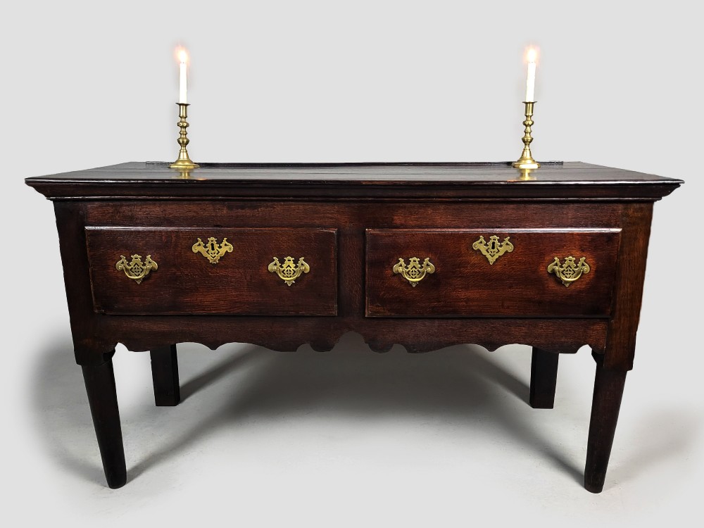 A George III dark oak dresser base featuring two drawers with ornate brass handles and escutcheons, a shaped apron, and tapered legs. View IV