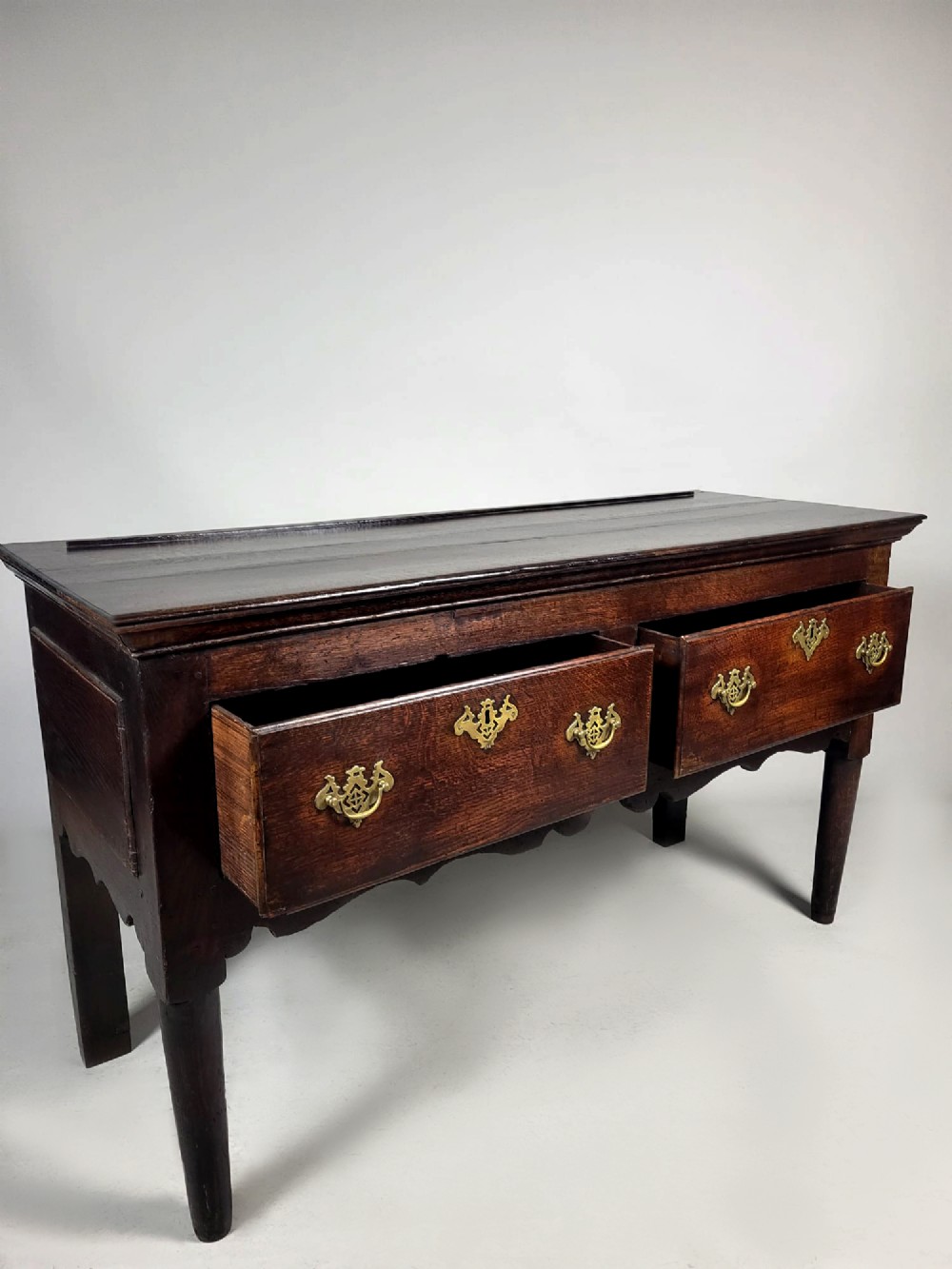 A George III dark oak dresser base featuring two drawers with ornate brass handles and escutcheons, a shaped apron, and tapered legs. View V