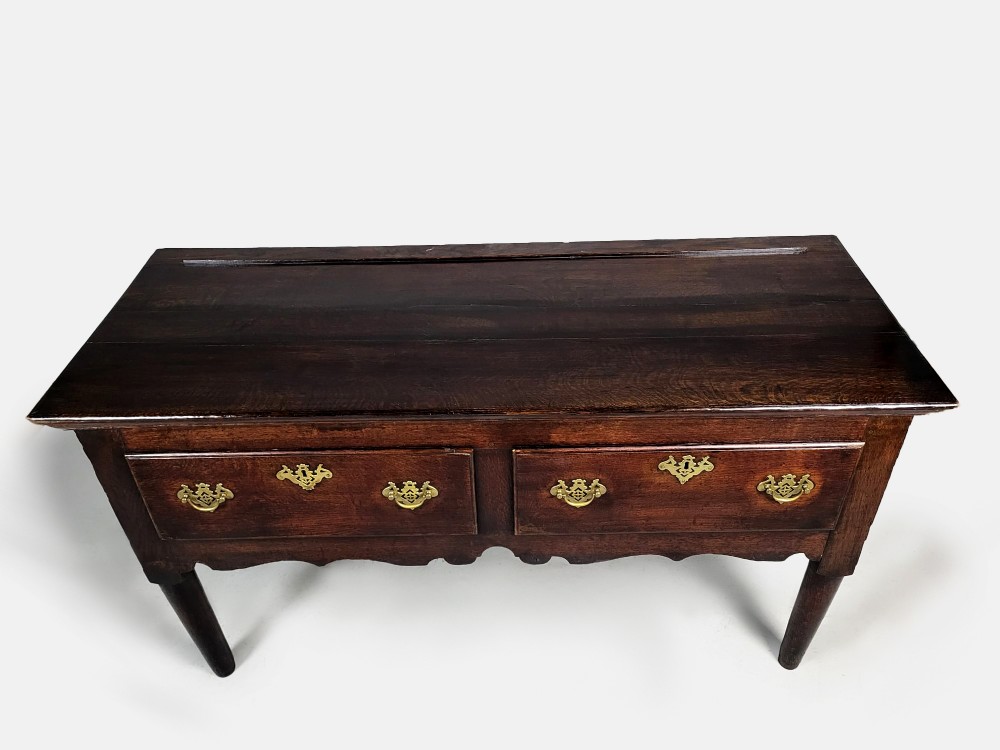 A George III dark oak dresser base featuring two drawers with ornate brass handles and escutcheons, a shaped apron, and tapered legs. View VI