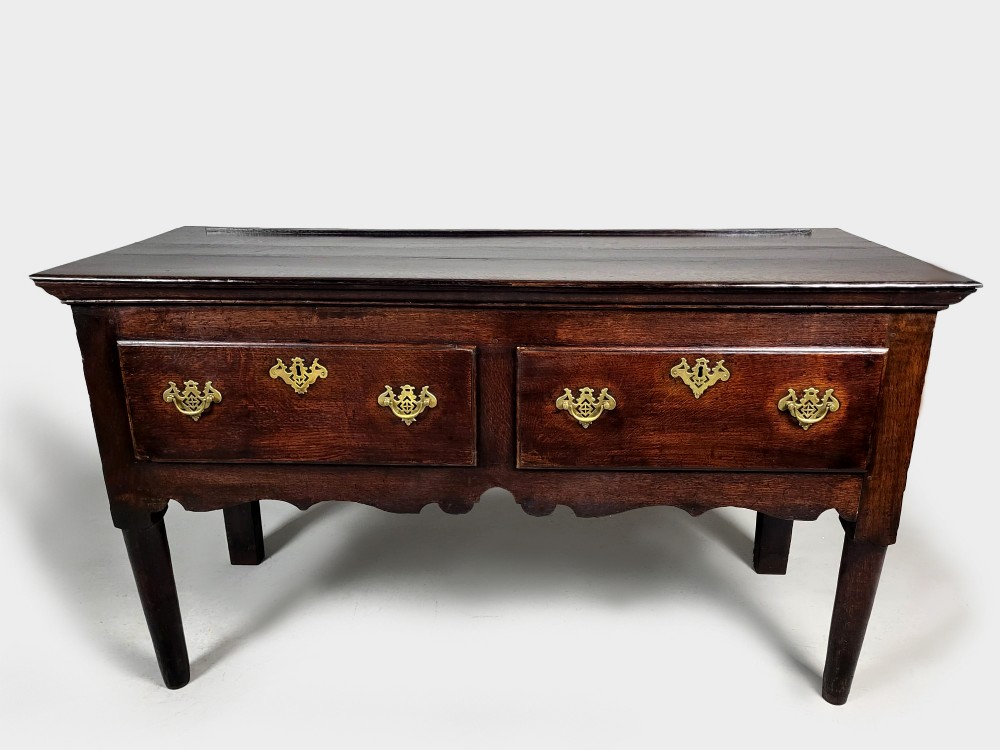 A George III dark oak dresser base featuring two drawers with ornate brass handles and escutcheons, a shaped apron, and tapered legs. View VII