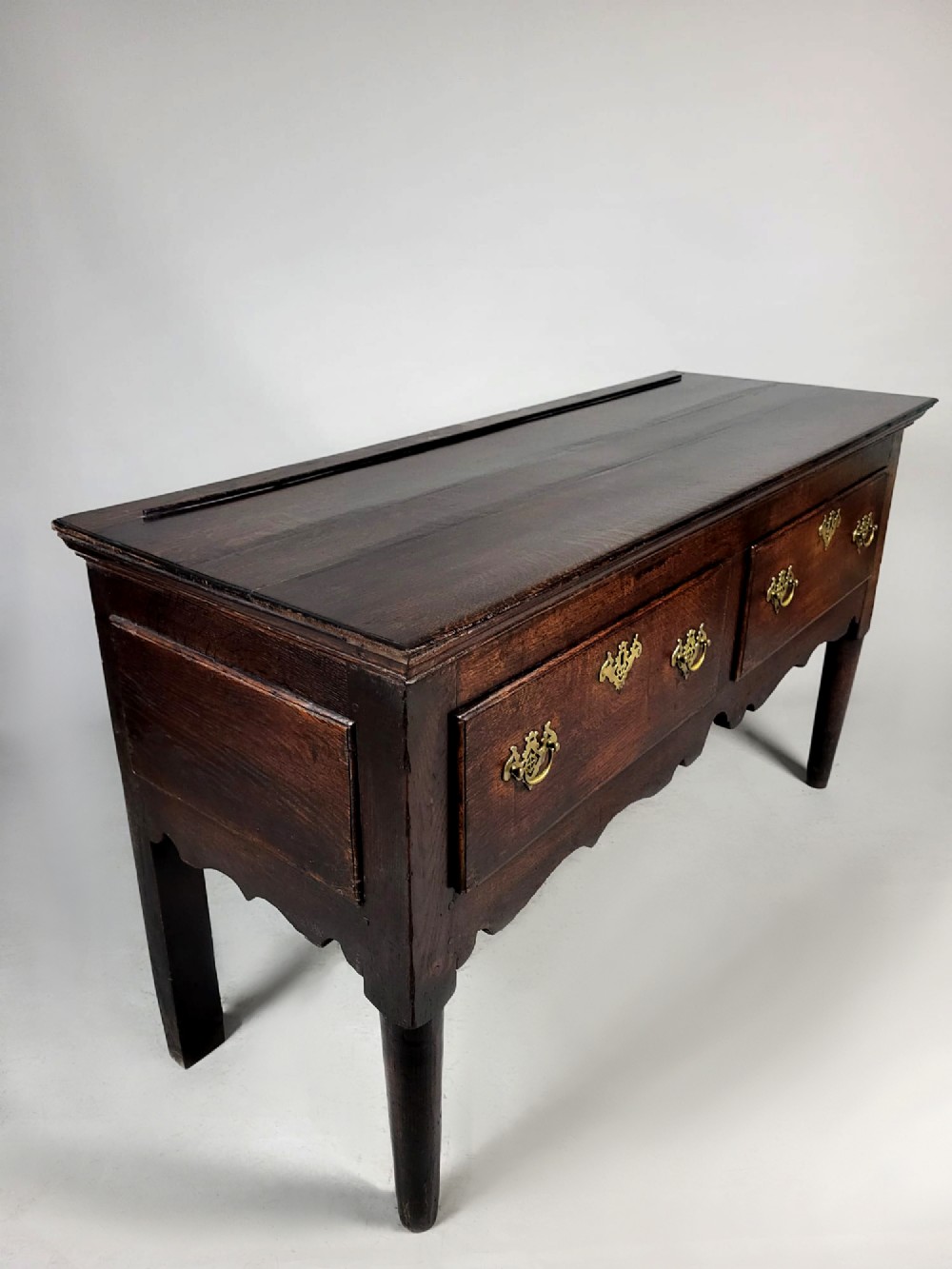 A George III dark oak dresser base featuring two drawers with ornate brass handles and escutcheons, a shaped apron, and tapered legs. View VIII