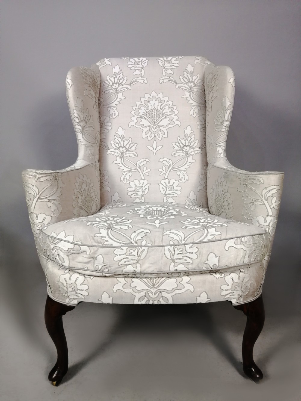 A Victorian wingback armchair upholstered in light cream floral damask fabric with dark carved walnut cabriole legs.