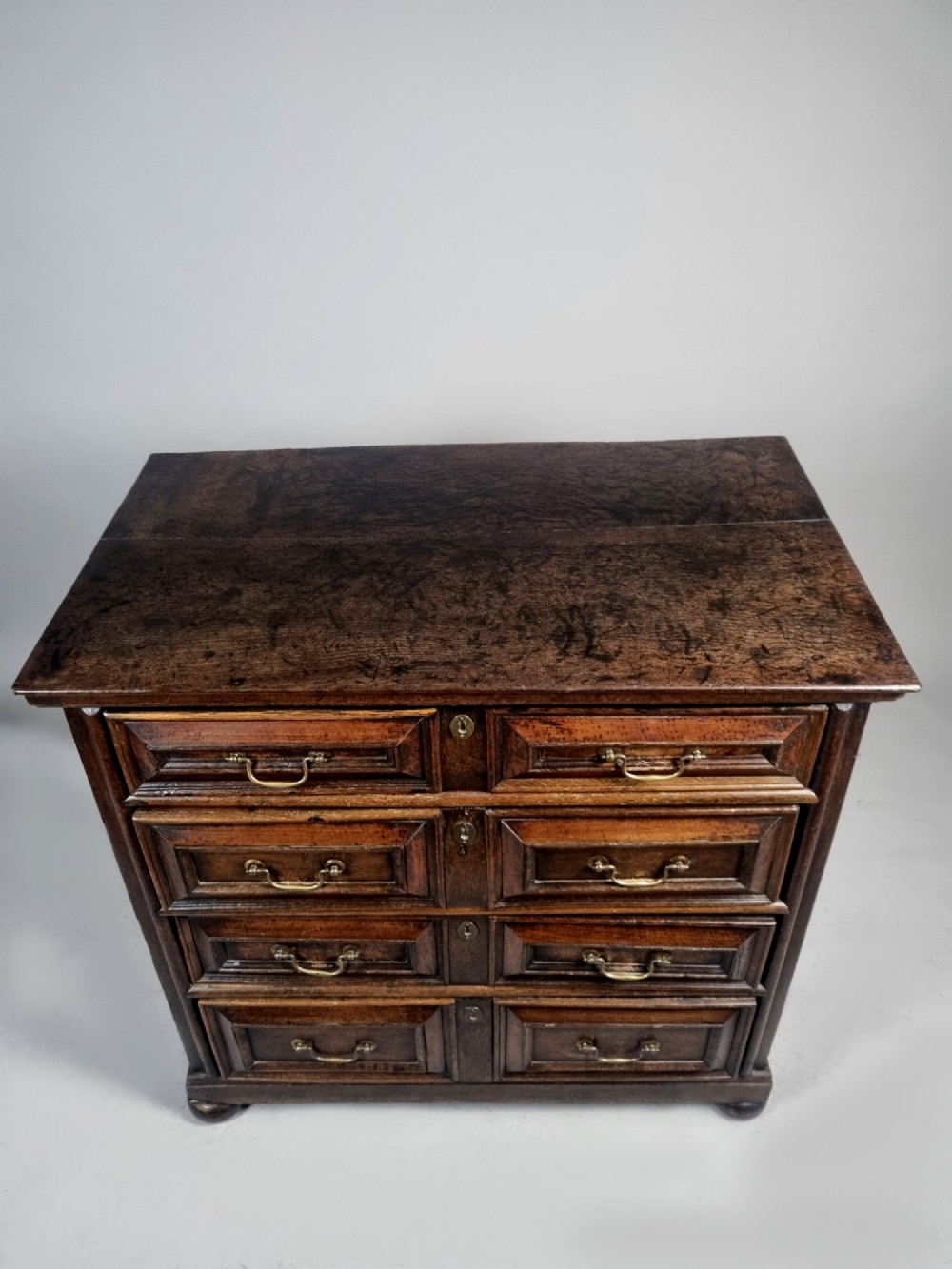 A seventeenth-century dark oak chest of drawers featuring geometric mitred drawer panels, brass swing handles, and turned bun feet. View IV