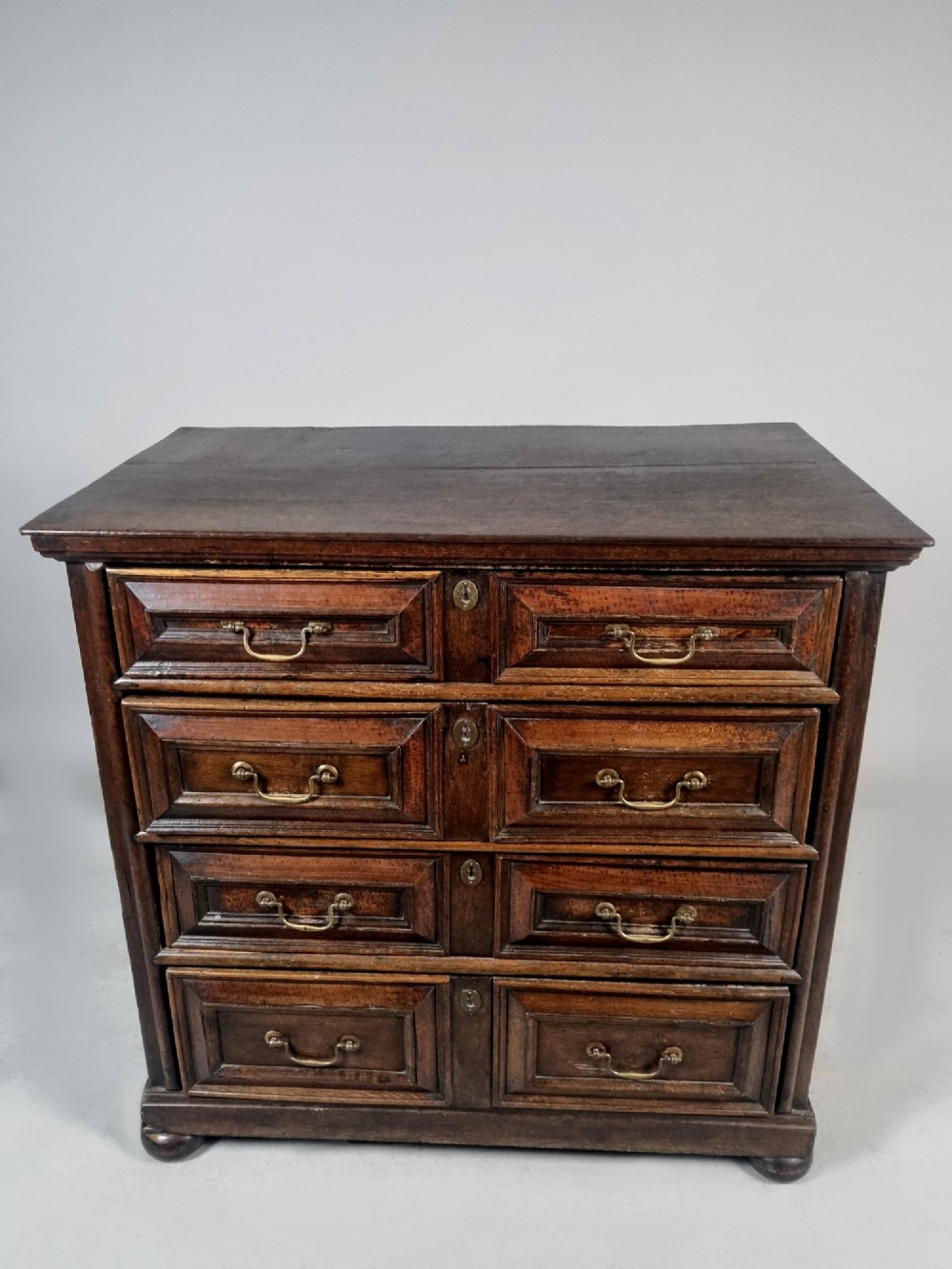 A seventeenth-century dark oak chest of drawers featuring geometric mitred drawer panels, brass swing handles, and turned bun feet. View V