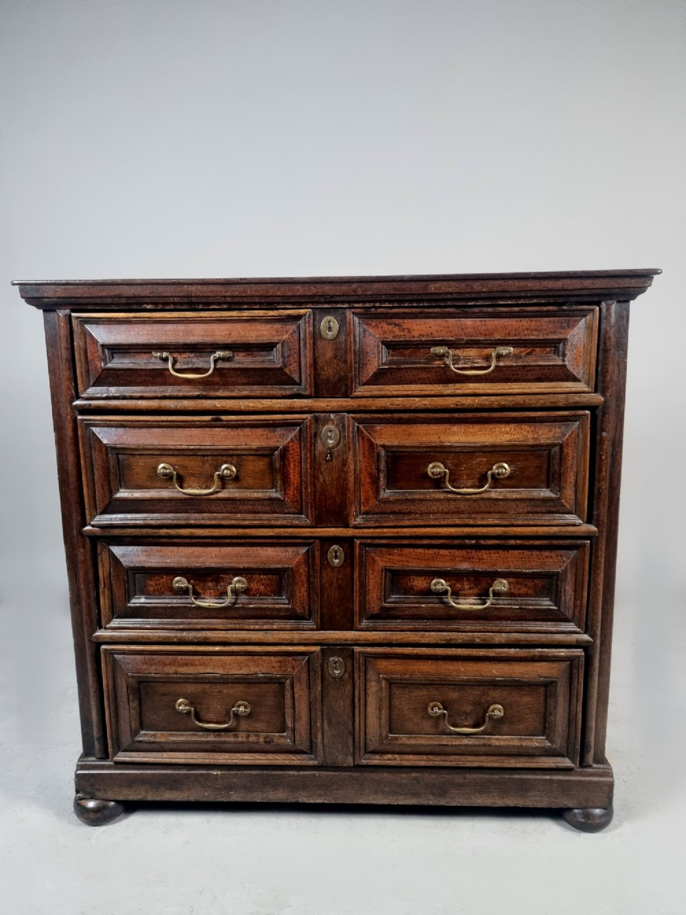A seventeenth-century dark oak chest of drawers featuring geometric mitred drawer panels, brass swing handles, and turned bun feet. View VI