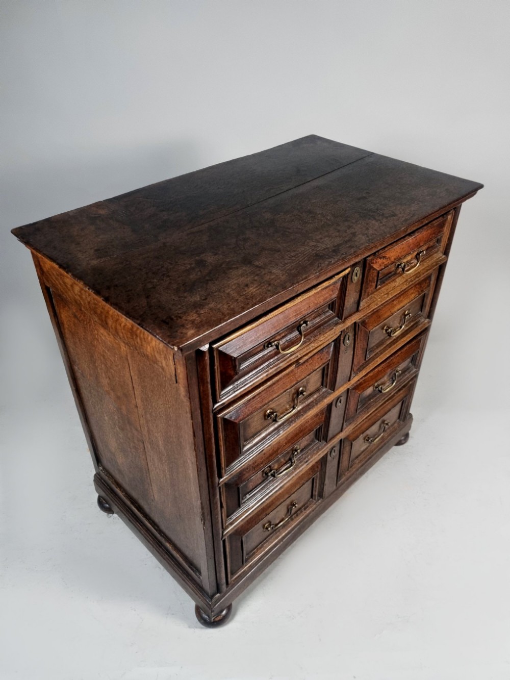 A seventeenth-century dark oak chest of drawers featuring geometric mitred drawer panels, brass swing handles, and turned bun feet. View VII