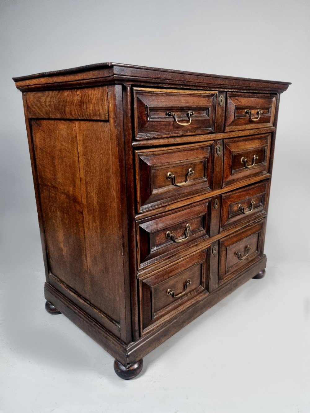 A seventeenth-century dark oak chest of drawers featuring geometric mitred drawer panels, brass swing handles, and turned bun feet. View VIII