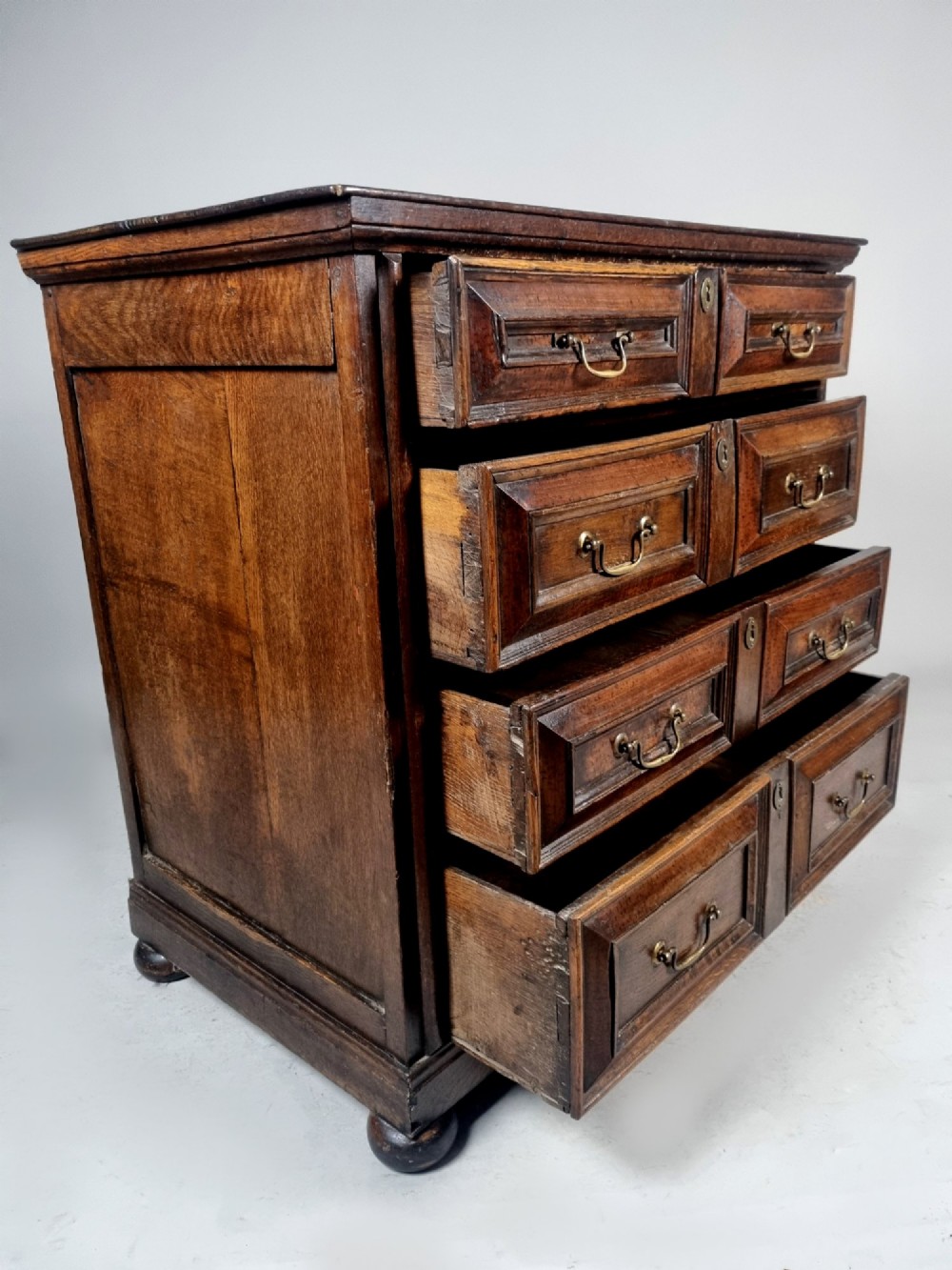 A seventeenth-century dark oak chest of drawers featuring geometric mitred drawer panels, brass swing handles, and turned bun feet. View IX