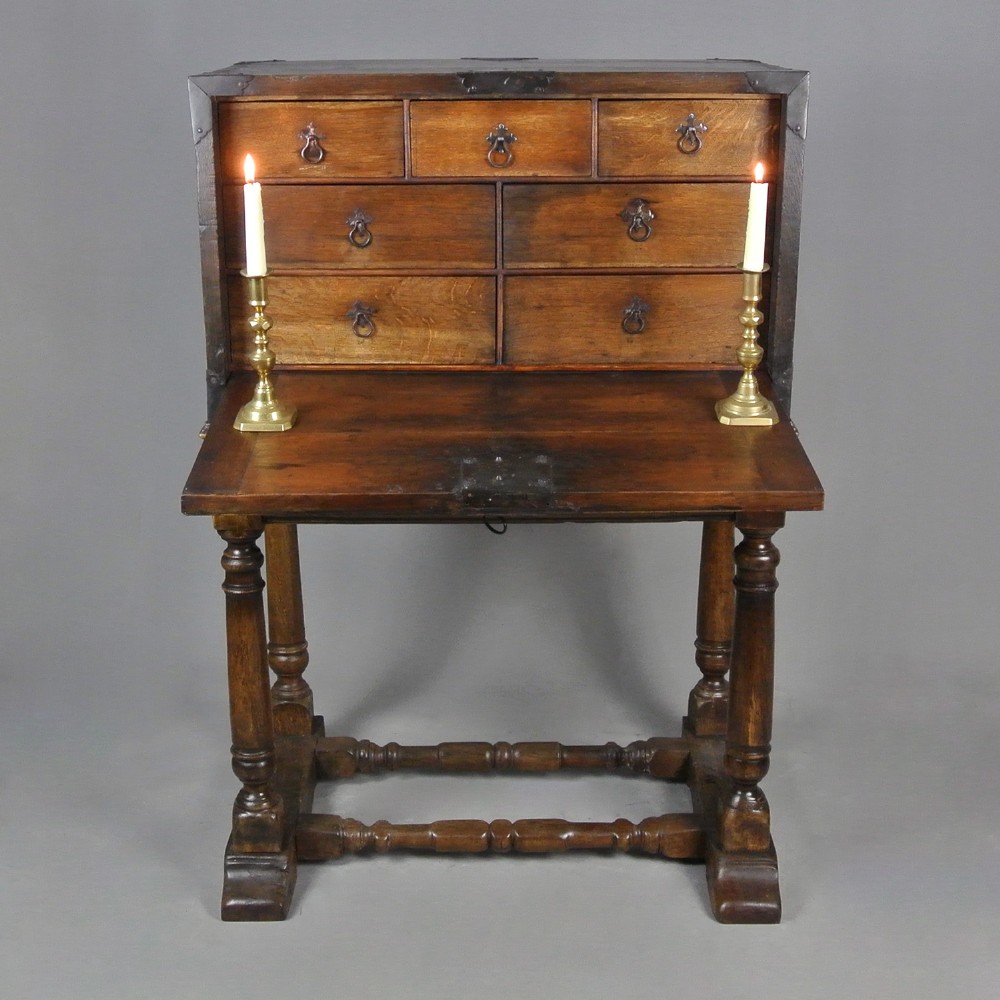 A seventeenth-century dark oak fall-front writing desk on a turned leg stand with interior drawers and wrought iron corner brackets.