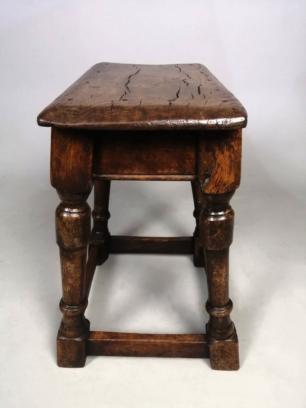 An antique dark brown oak joint stool with an elm top, turned legs, and perimeter box stretchers. View II