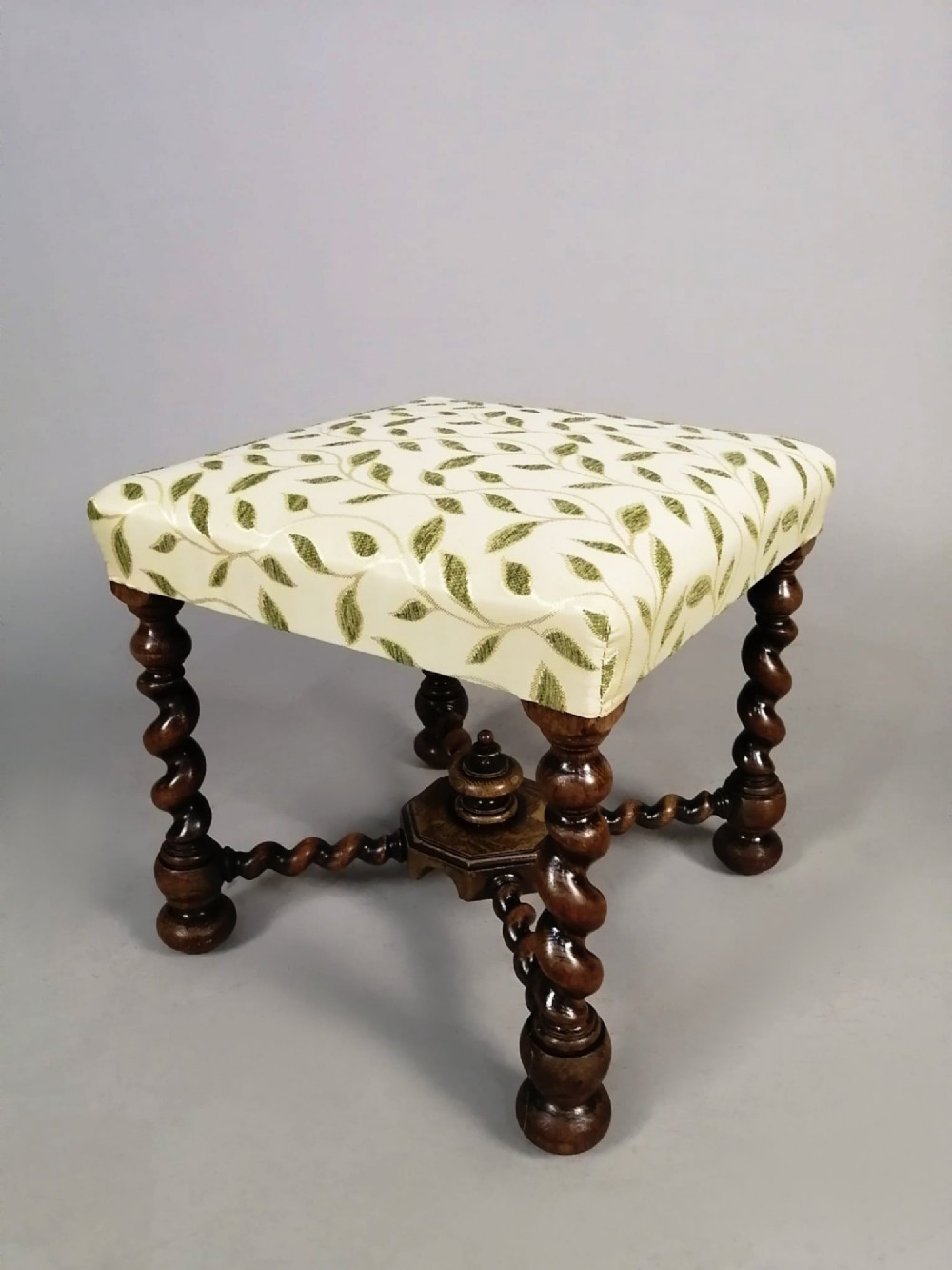 A square dark walnut stool featuring spiral barley twist legs, a central finial stretcher, and cream upholstery with embroidered green leaf motifs.