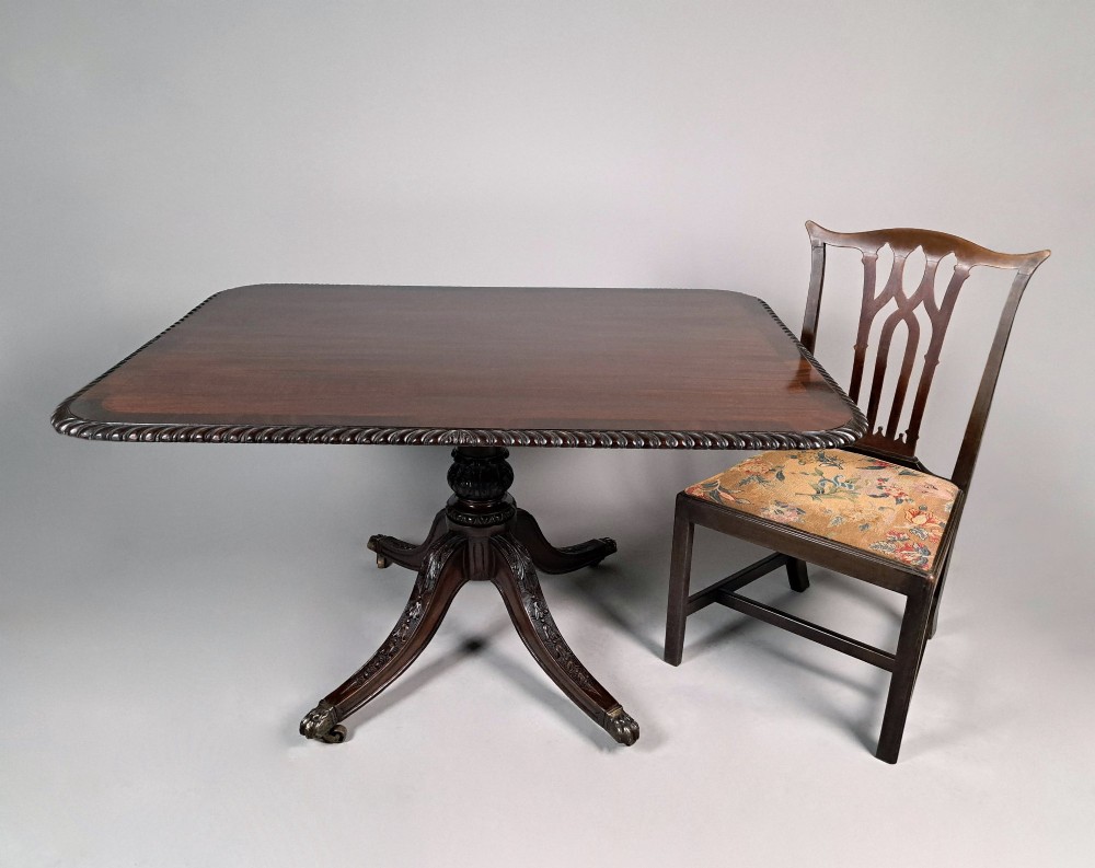 A George III mahogany dining table featuring a crossbanded top with gadrooned edging, supported on a carved pedestal with four splayed legs ending in brass castors. View VI