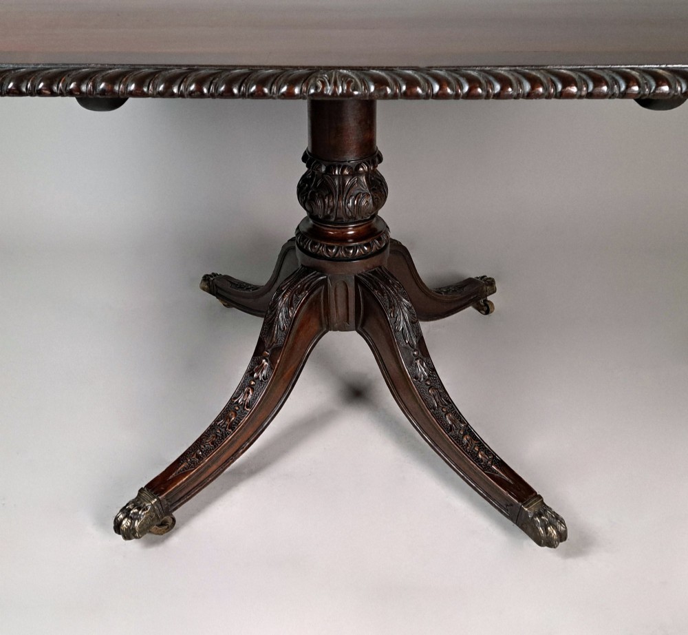 A George III mahogany dining table featuring a crossbanded top with gadrooned edging, supported on a carved pedestal with four splayed legs ending in brass castors. View VII