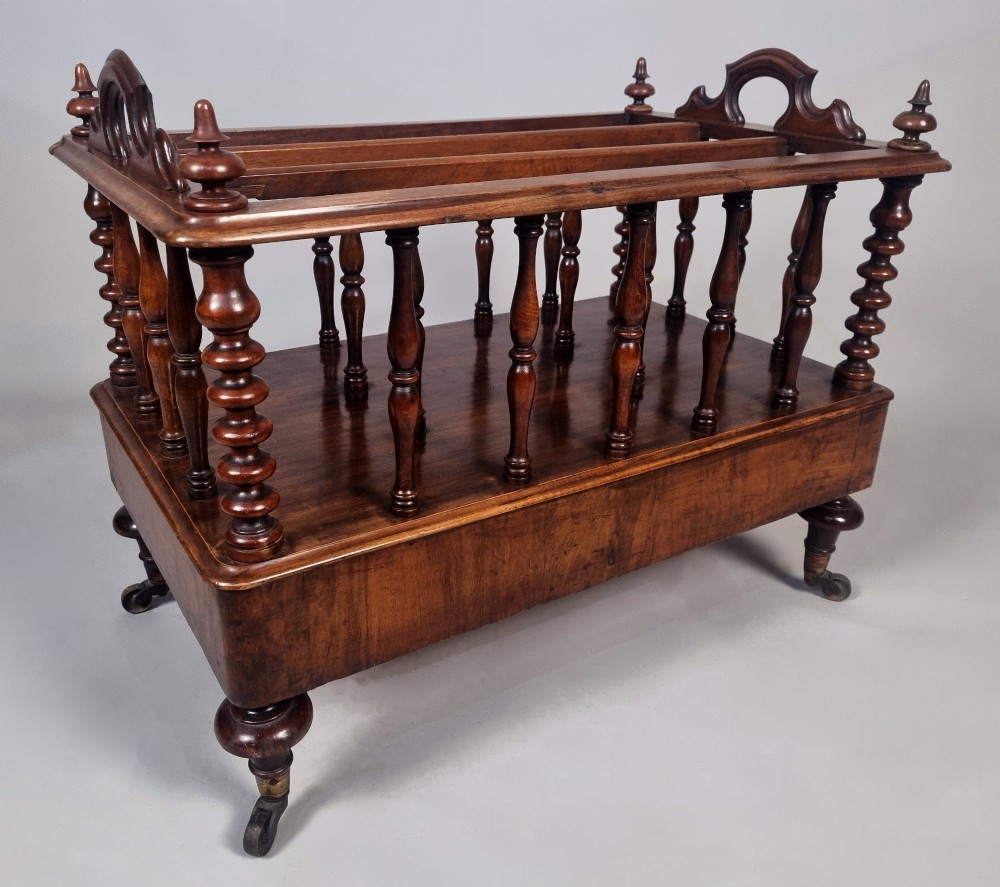 A Victorian walnut Canterbury featuring vertical spindle dividers, a lower drawer base, carved side handles, and turned feet with brass castors.
