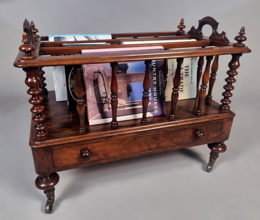 A Victorian walnut Canterbury featuring vertical spindle dividers, a lower drawer base, carved side handles, and turned feet with brass castors. View III