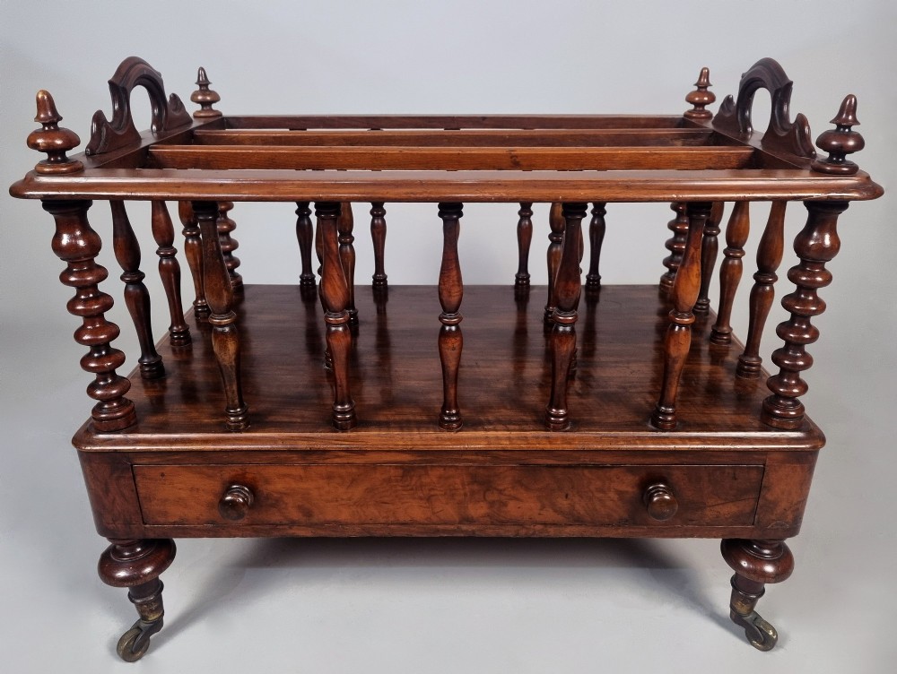 A Victorian walnut Canterbury featuring vertical spindle dividers, a lower drawer base, carved side handles, and turned feet with brass castors. View IV