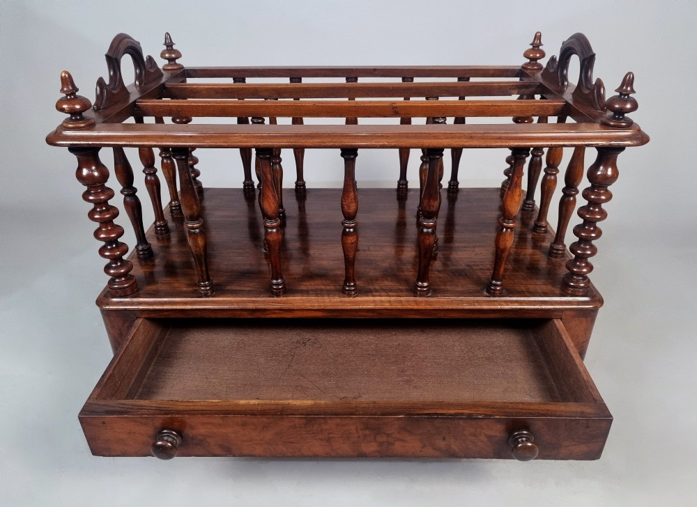 A Victorian walnut Canterbury featuring vertical spindle dividers, a lower drawer base, carved side handles, and turned feet with brass castors. View V