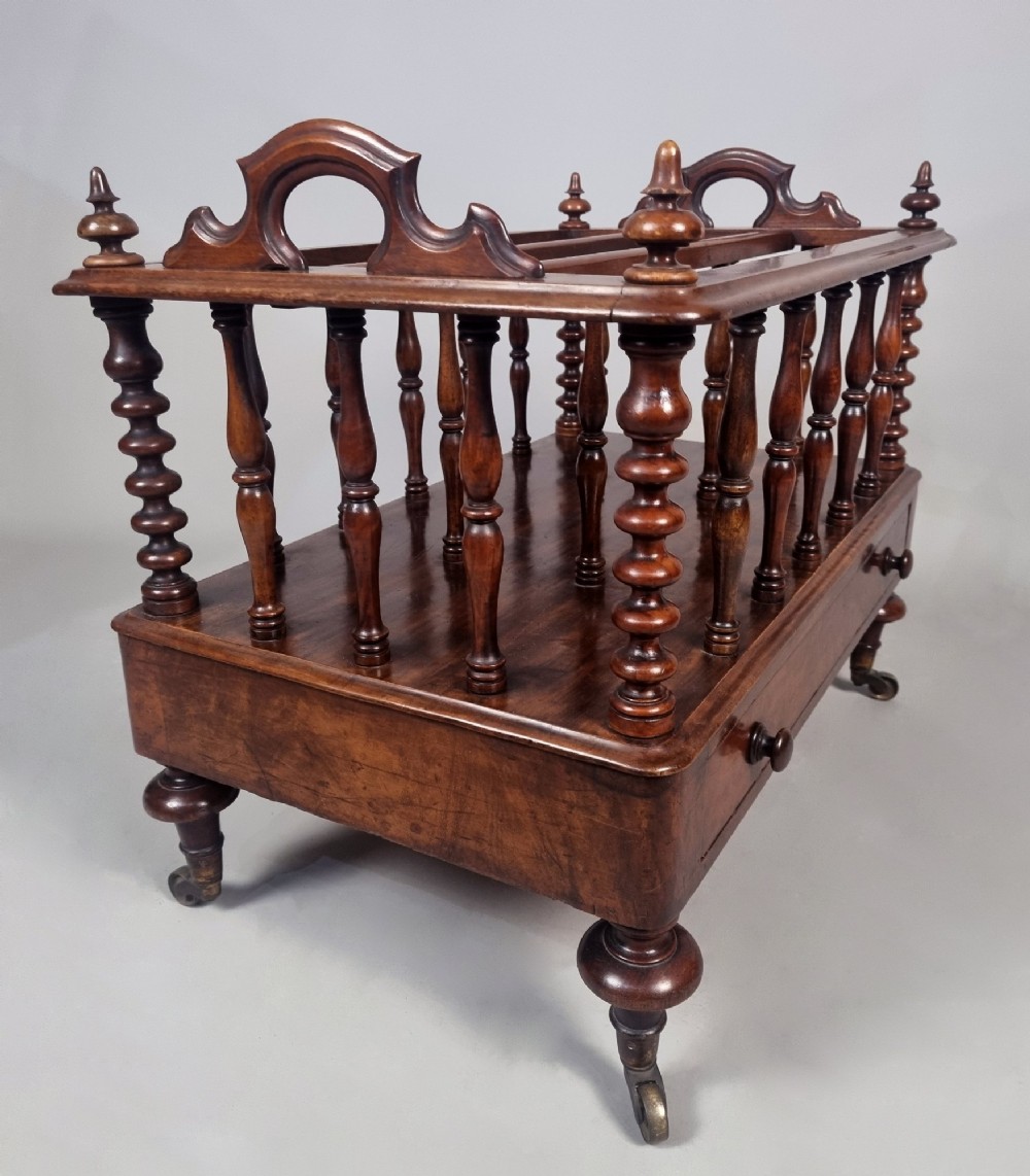 A Victorian walnut Canterbury featuring vertical spindle dividers, a lower drawer base, carved side handles, and turned feet with brass castors. View VI
