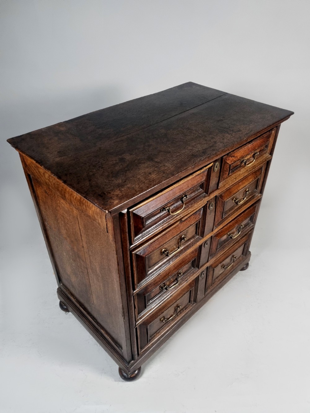 A seventeenth-century Charles II solid oak chest of drawers featuring geometric panelled drawer fronts, brass drop handles, and bun feet. View VII
