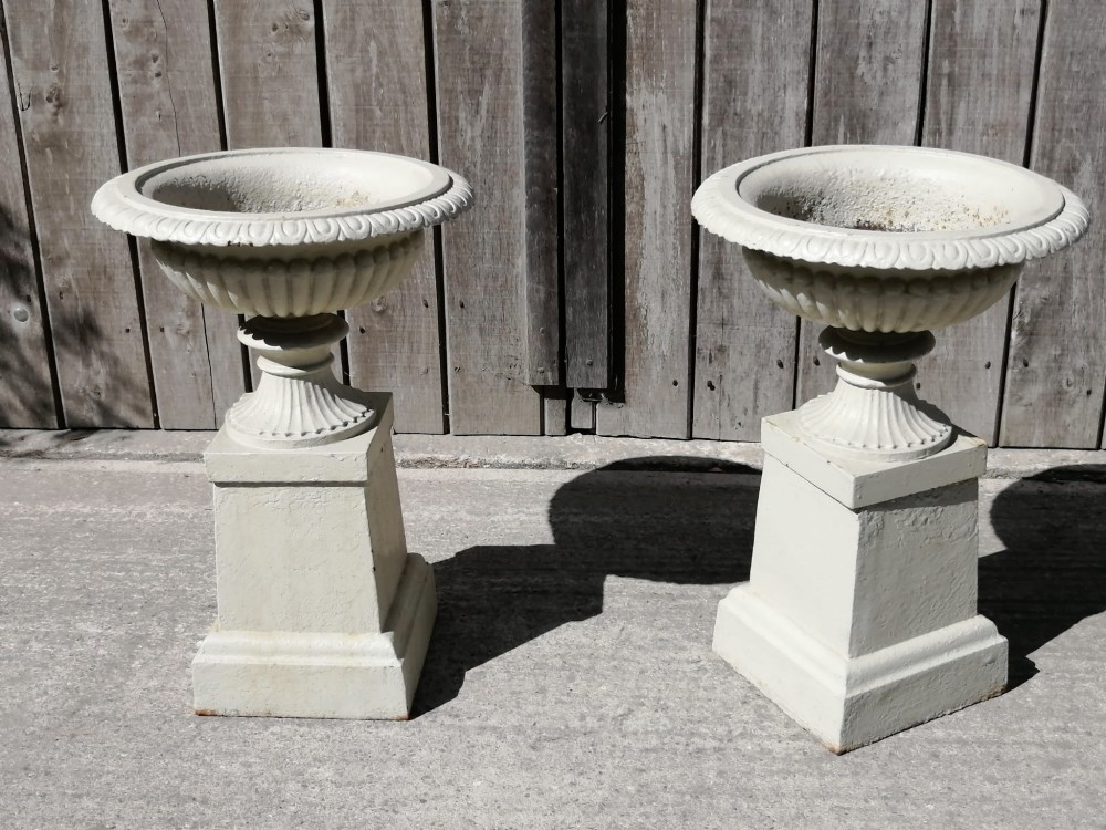 A pair of off-white painted cast iron garden urns featuring fluted rims and bodies set upon square plinths. View II