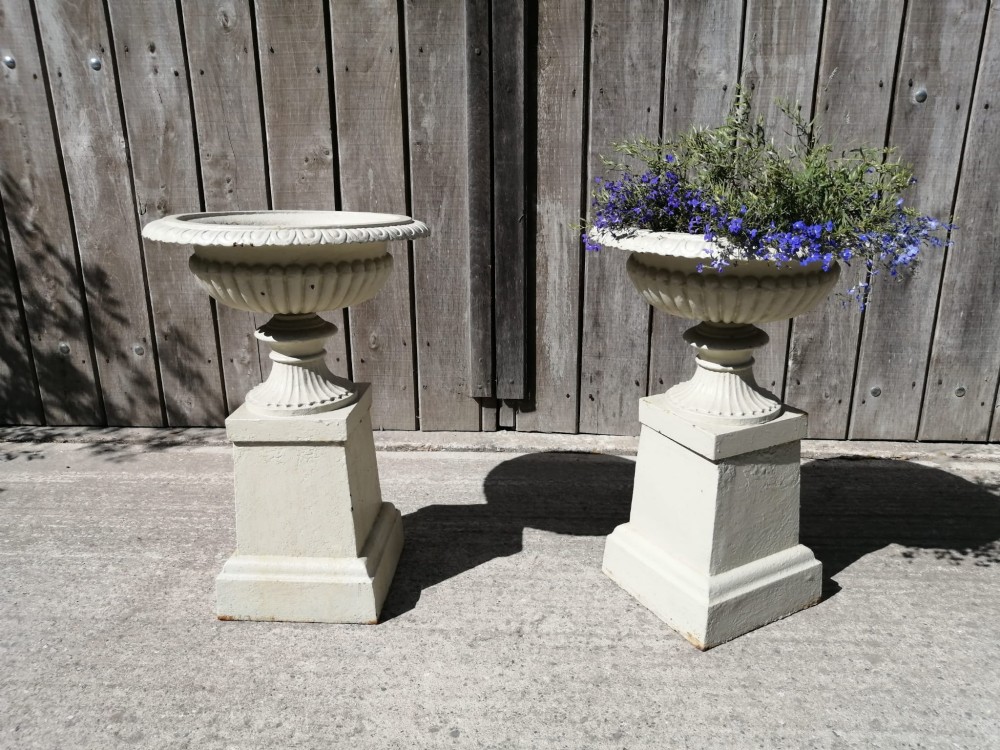 A pair of off-white painted cast iron garden urns featuring fluted rims and bodies set upon square plinths. View VI