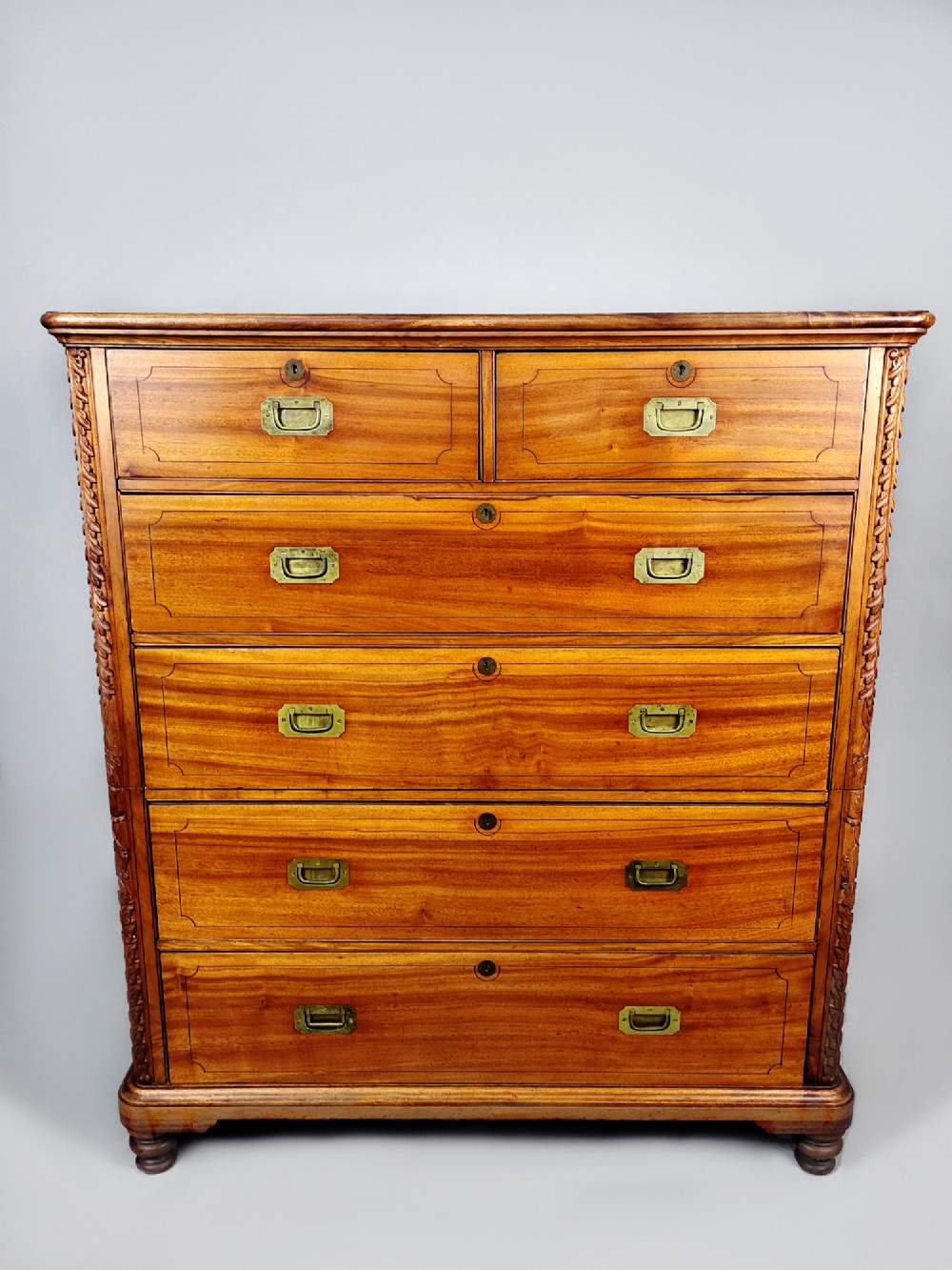 A nineteenth-century teak campaign chest of drawers featuring flush brass handles, keyholes, two short over three long drawers, and carved side pilasters.