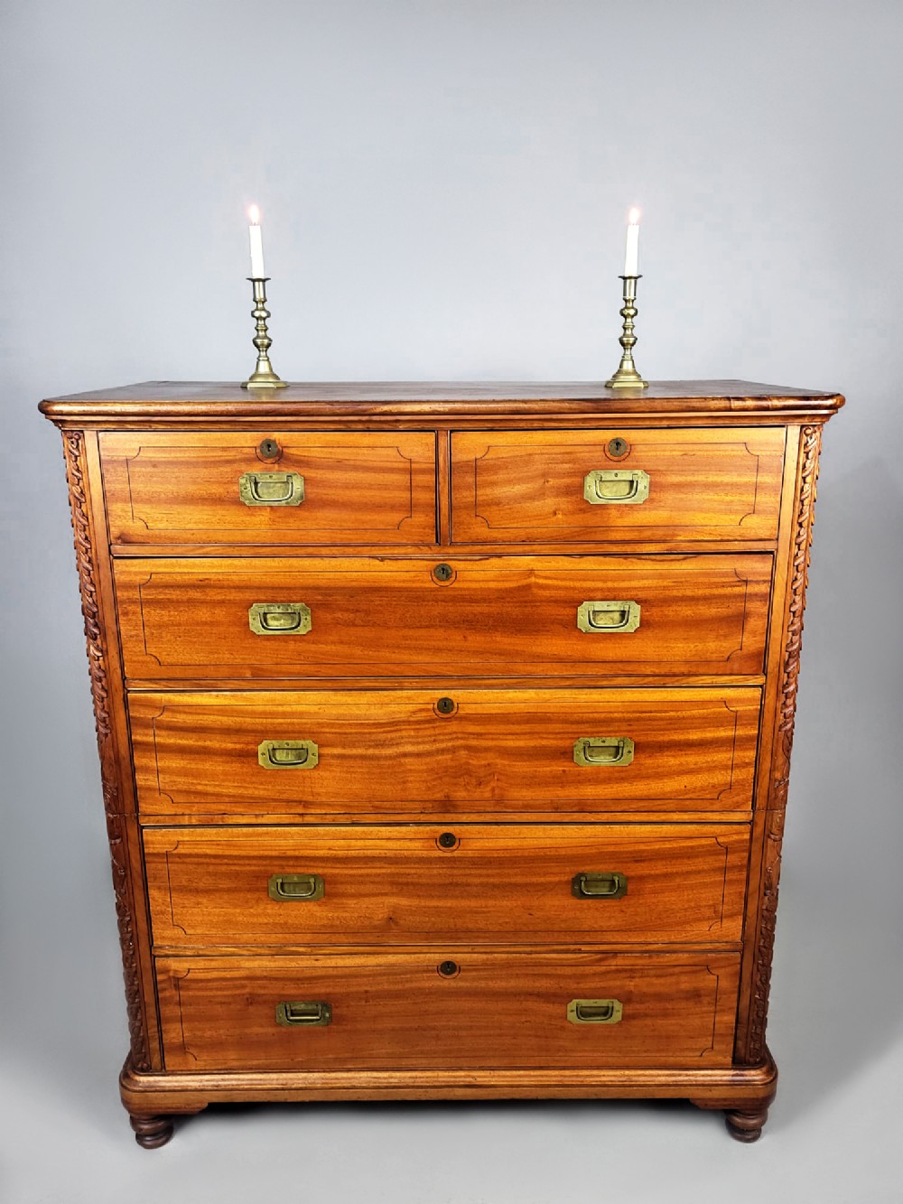 A nineteenth-century teak campaign chest of drawers featuring flush brass handles, keyholes, two short over three long drawers, and carved side pilasters. View IV