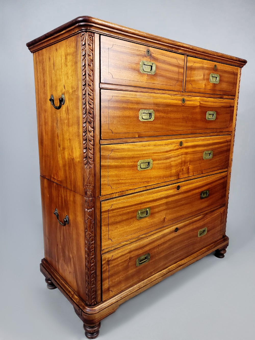 A nineteenth-century teak campaign chest of drawers featuring flush brass handles, keyholes, two short over three long drawers, and carved side pilasters. View V