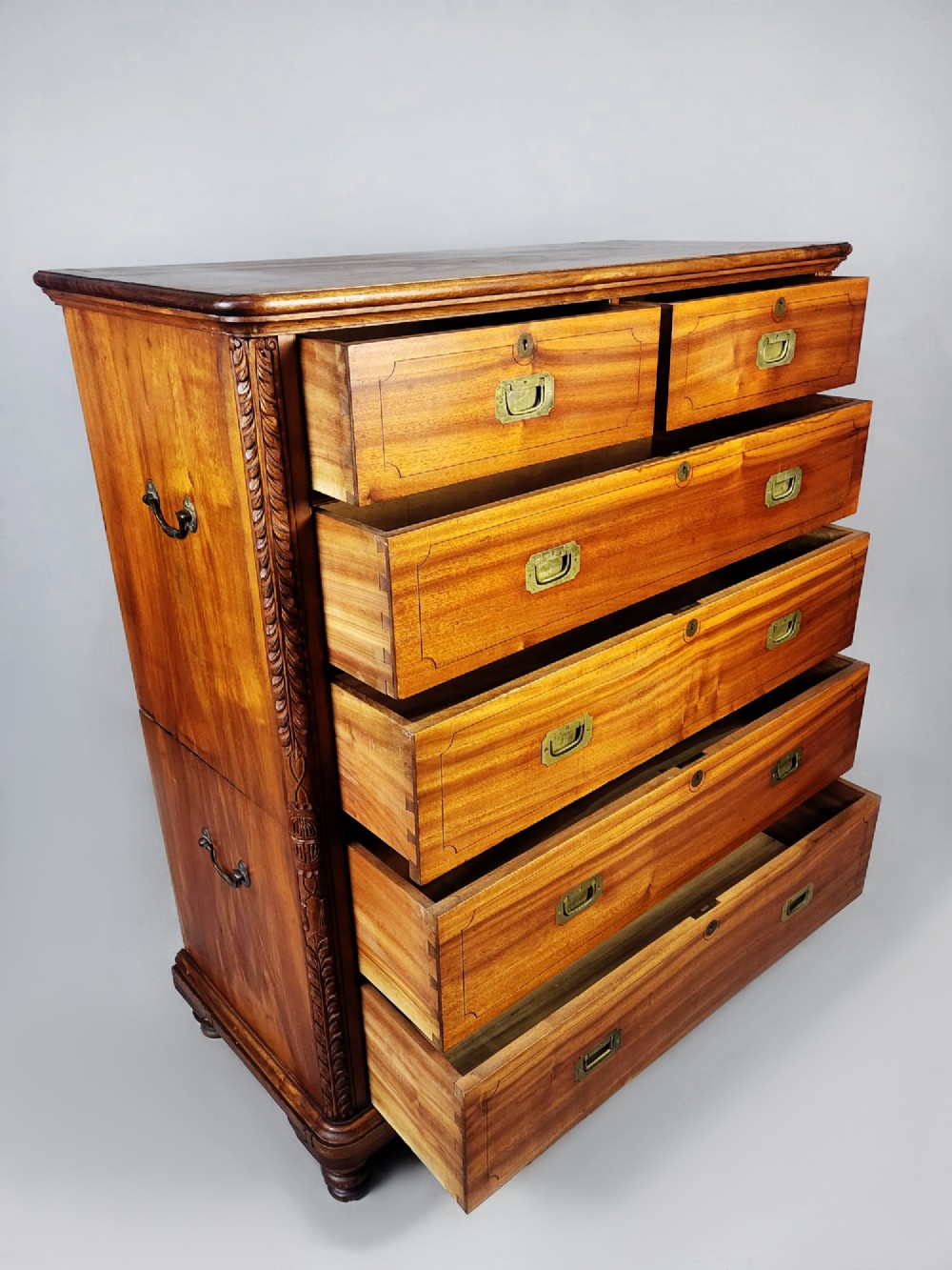 A nineteenth-century teak campaign chest of drawers featuring flush brass handles, keyholes, two short over three long drawers, and carved side pilasters. View VI