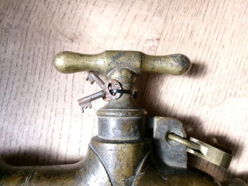 A heavy Victorian brass barrel tap featuring a tapered shaft, curved spout, T-bar handle, and an integrated locking loop mechanism. View IV
