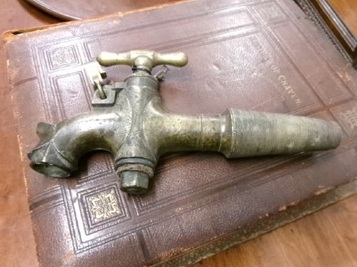 A heavy Victorian brass barrel tap featuring a tapered shaft, curved spout, T-bar handle, and an integrated locking loop mechanism. View VII