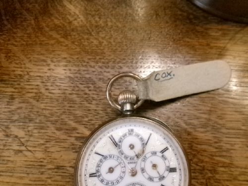 A gunmetal calendar moonphase pocket watch with an enamel dial displayed upright inside a turned wooden holder. View VII