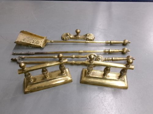 A brass fire companion set featuring two baluster-turned fire dogs, an iron rest, a pierced shovel, a poker, and tongs laid out on a surface. View II
