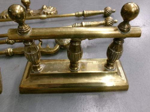 A brass fire companion set featuring two baluster-turned fire dogs, an iron rest, a pierced shovel, a poker, and tongs laid out on a surface. View III