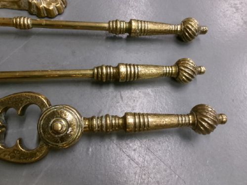 A brass fire companion set featuring two baluster-turned fire dogs, an iron rest, a pierced shovel, a poker, and tongs laid out on a surface. View IV