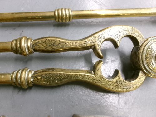A brass fire companion set featuring two baluster-turned fire dogs, an iron rest, a pierced shovel, a poker, and tongs laid out on a surface. View V