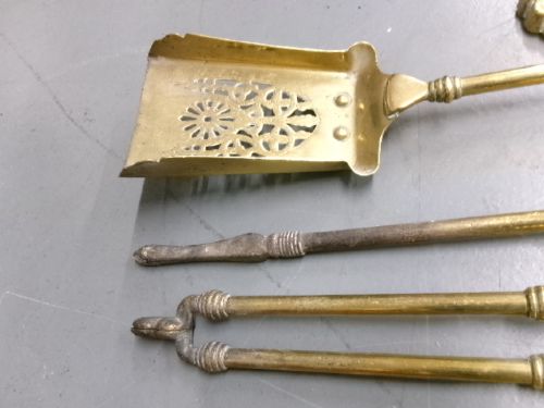 A brass fire companion set featuring two baluster-turned fire dogs, an iron rest, a pierced shovel, a poker, and tongs laid out on a surface. View VII