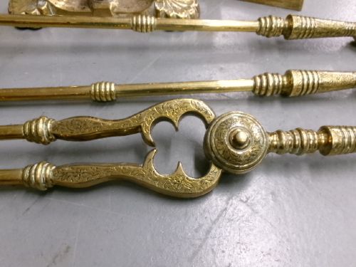 A brass fire companion set featuring two baluster-turned fire dogs, an iron rest, a pierced shovel, a poker, and tongs laid out on a surface. View IX