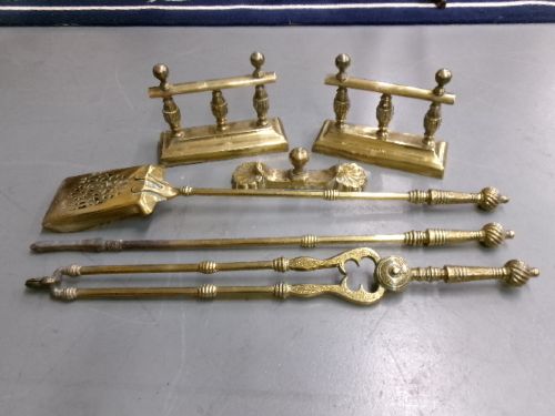 A brass fire companion set featuring two baluster-turned fire dogs, an iron rest, a pierced shovel, a poker, and tongs laid out on a surface. View X