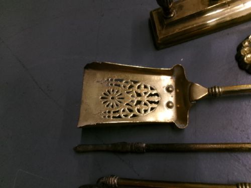 A brass fire companion set featuring two baluster-turned fire dogs, an iron rest, a pierced shovel, a poker, and tongs laid out on a surface. View XII