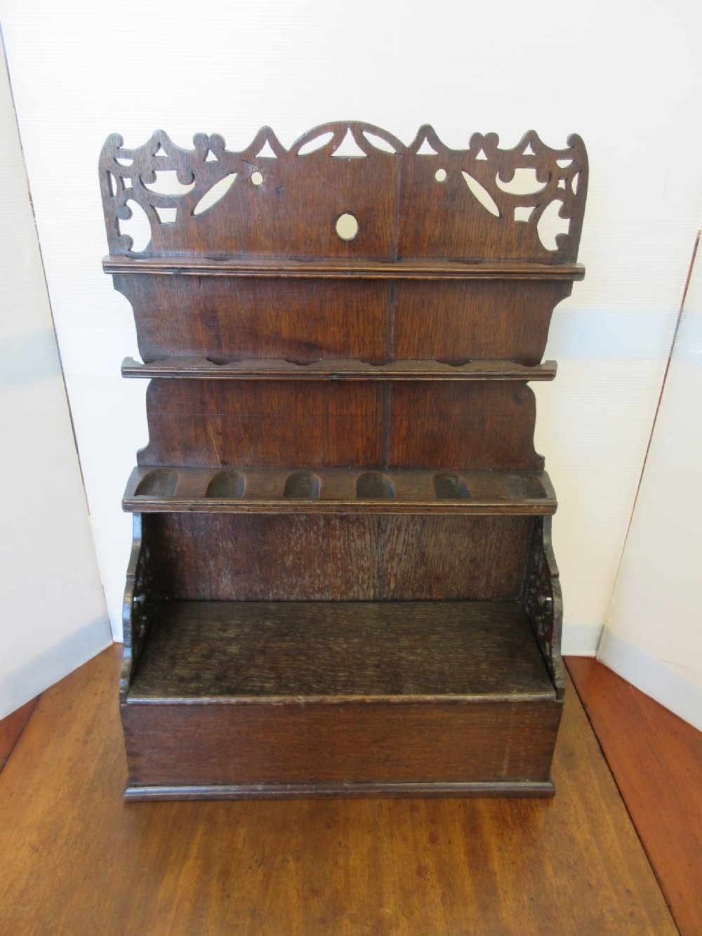 A dark brown carved oak wall rack features multiple tiered shelves, slots for pipes, and an ornately pierced crest at the top.